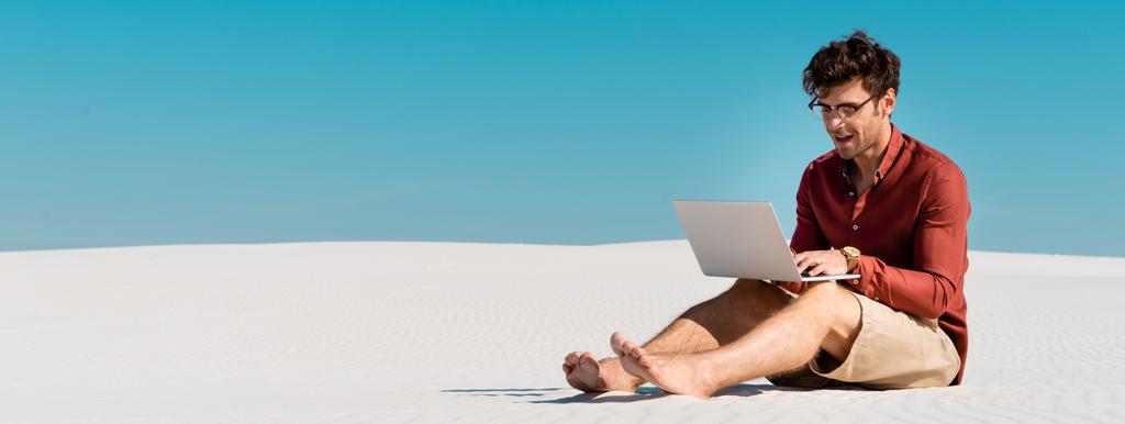Genç serbest çalışan kumlu sahilde açık mavi gökyüzüne karşı dizüstü bilgisayar kullanıyor, panoramik çekim - Fotoğraf, Görsel