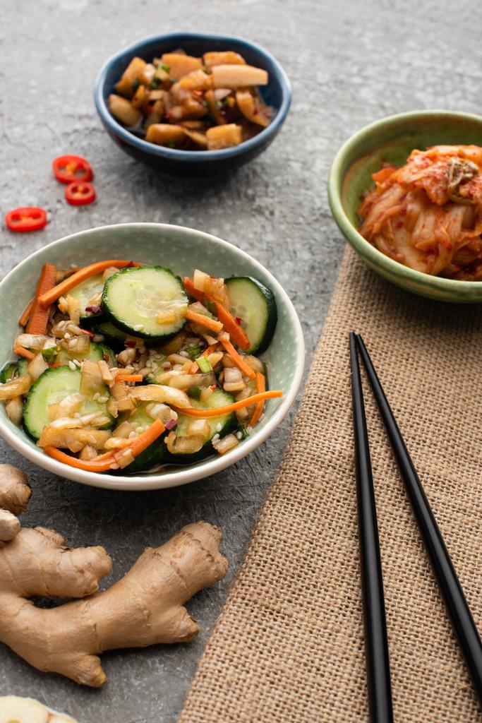 selektiver Fokus von Schalen mit schmackhaften Kimchi in der Nähe von Essstäbchen und Ingwer auf Betonoberfläche - Foto, Bild