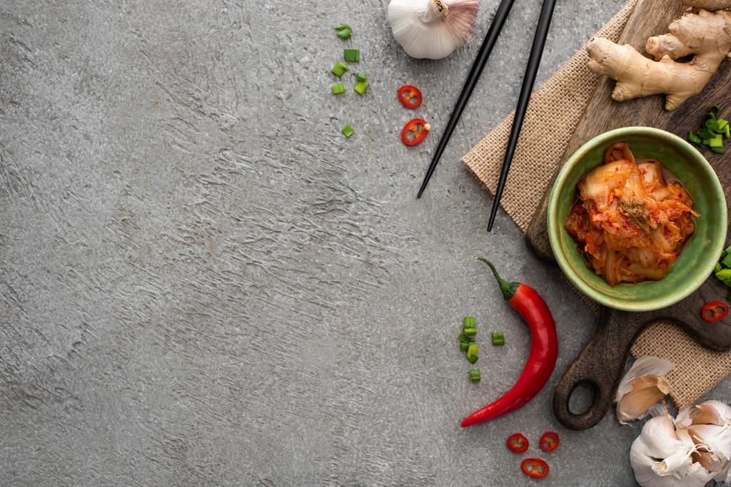 bovenaanzicht van kom met kimchi op snijplank bij eetstokjes, gember, chili peper en knoflook op betonnen ondergrond - Foto, afbeelding