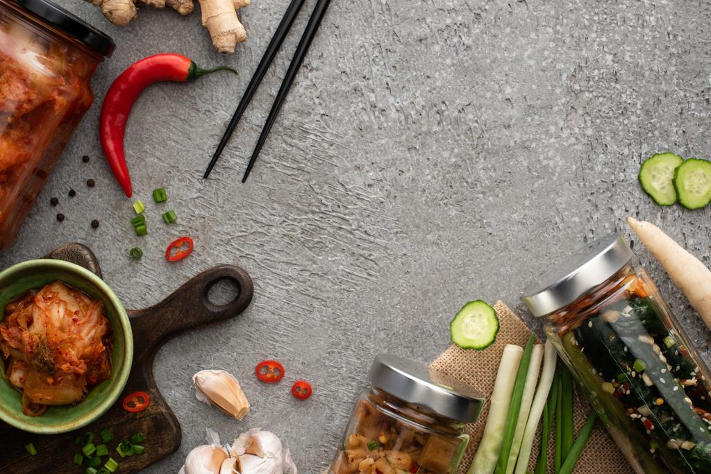 felső kilátás ízletes kimchi üvegek és tál közelében evőpálcikák, zöld hagyma, gyömbér, fokhagyma és chili paprika beton felületen - Fotó, kép