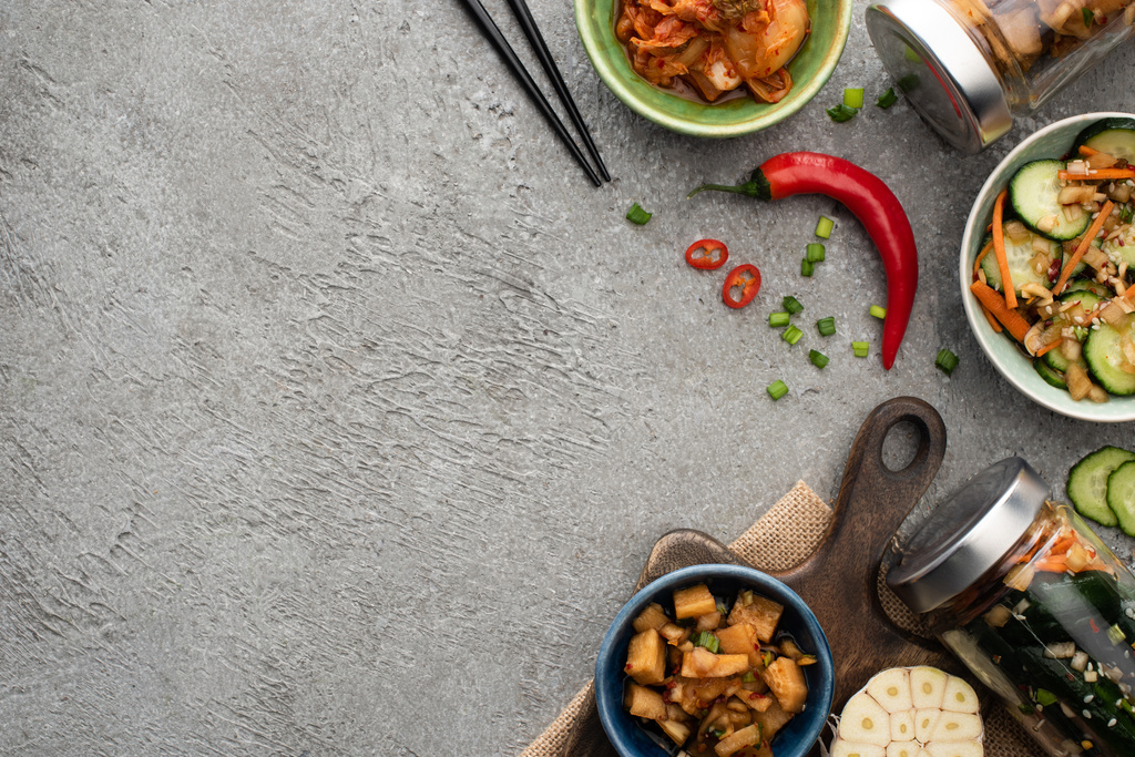 Blick von oben auf schmackhafte Kimchi in Schüsseln und Gläsern, neben Essstäbchen und Chilipfeffer auf Betonoberfläche - Foto, Bild
