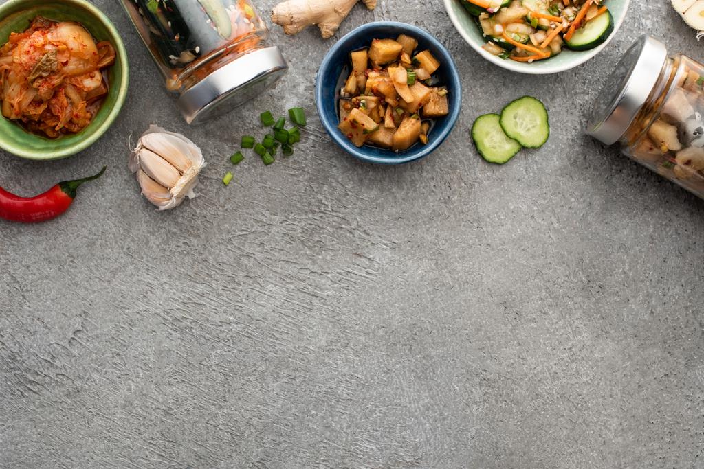 bovenaanzicht van smakelijke kimchi in kommen en potten bij eetstokjes, knoflook en chili peper op betonnen ondergrond - Foto, afbeelding