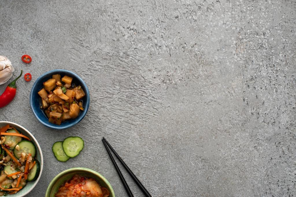 vista superior de cuencos con delicioso kimchi cerca de ajo, pepino en rodajas, chile y palillos en la superficie de hormigón
 - Foto, Imagen