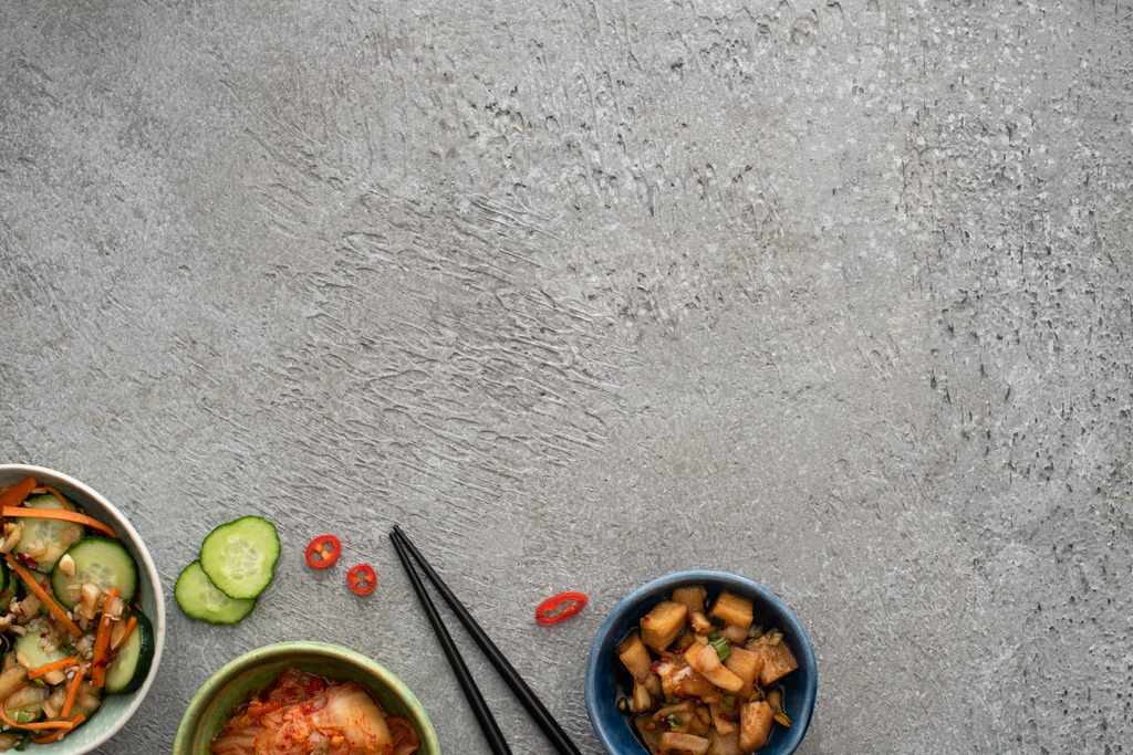 Schüsseln mit köstlichen Kimchi in der Nähe von Gurkenscheiben und Essstäbchen auf Betonoberfläche - Foto, Bild