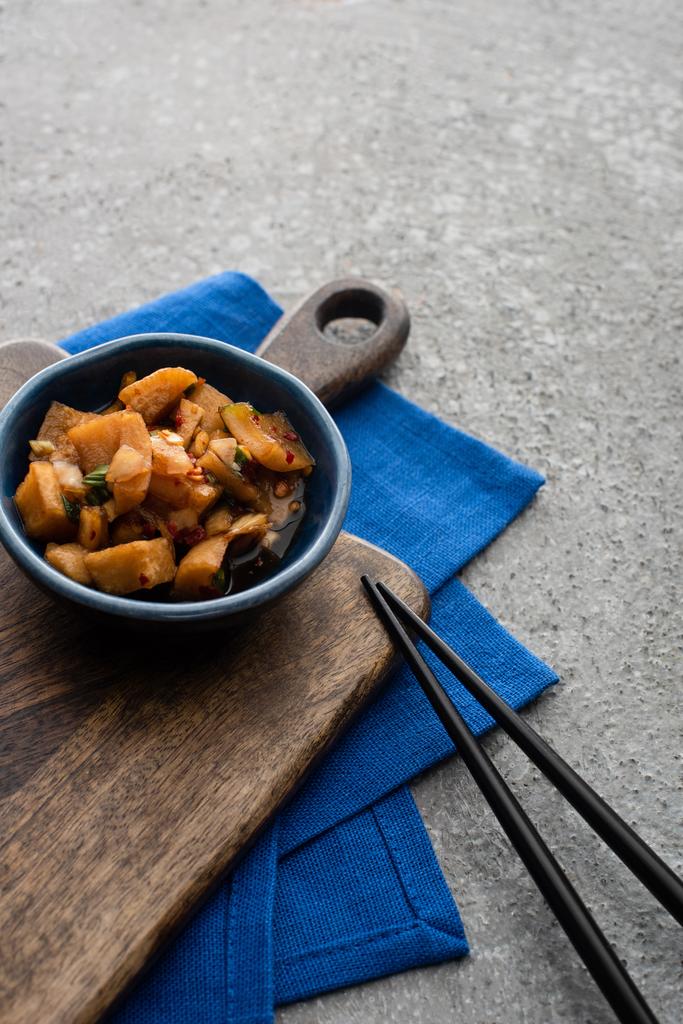bowl with deicious kimchi on wooden cutting board and blue cloth near chopsticks on concrete surface - Photo, Image