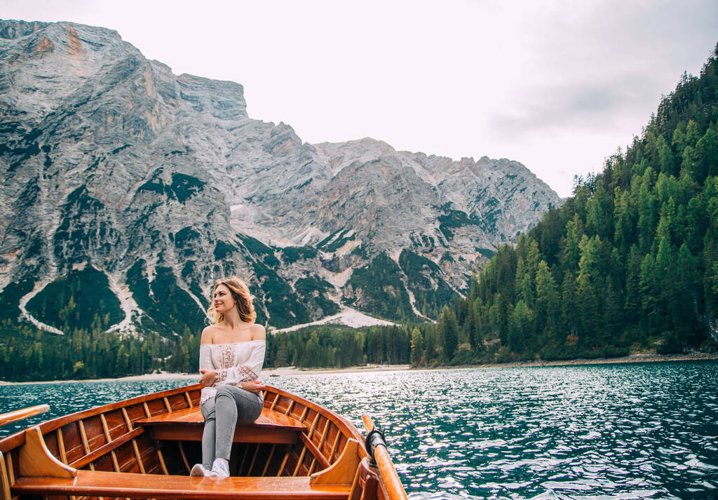 Beautiful redhaired girl clothes in white silk sexy blouse long sleeves bare shoulders gray trousers sitting in boat. Tourist enjoy view alpine lake of Braies. Backdrop Italian mountains green forest - Fotoğraf, Görsel