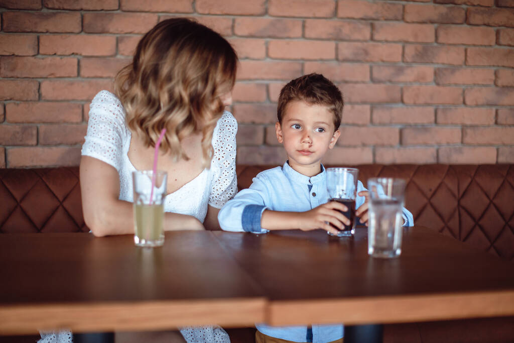 Χαριτωμένο αγόρι πίνει αναψυκτικό στο καφέ με τη μαμά του δίπλα του. Ανθυγιεινές παιδικές συνήθειες - Φωτογραφία, εικόνα