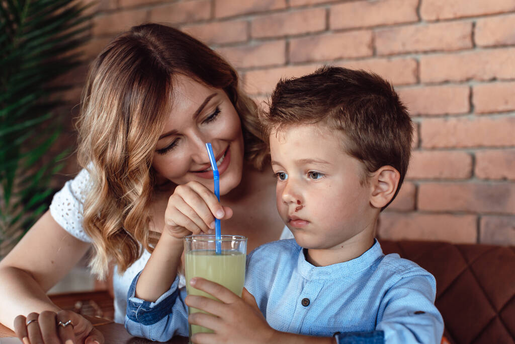 Aranyos fiú üdítőt iszik a kávézóban mellette az anyjával, és szeretettel nézi. - Fotó, kép