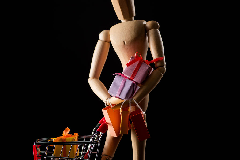 Ξύλινη κούκλα με πολύχρωμα δώρα και καλάθι αγορών που απομονώνονται σε μαύρο - Φωτογραφία, εικόνα