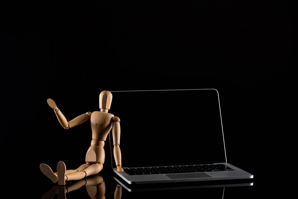 Ξύλινη κούκλα σε καθιστή θέση με απλωμένο χέρι κοντά στο laptop σε μαύρο  - Φωτογραφία, εικόνα