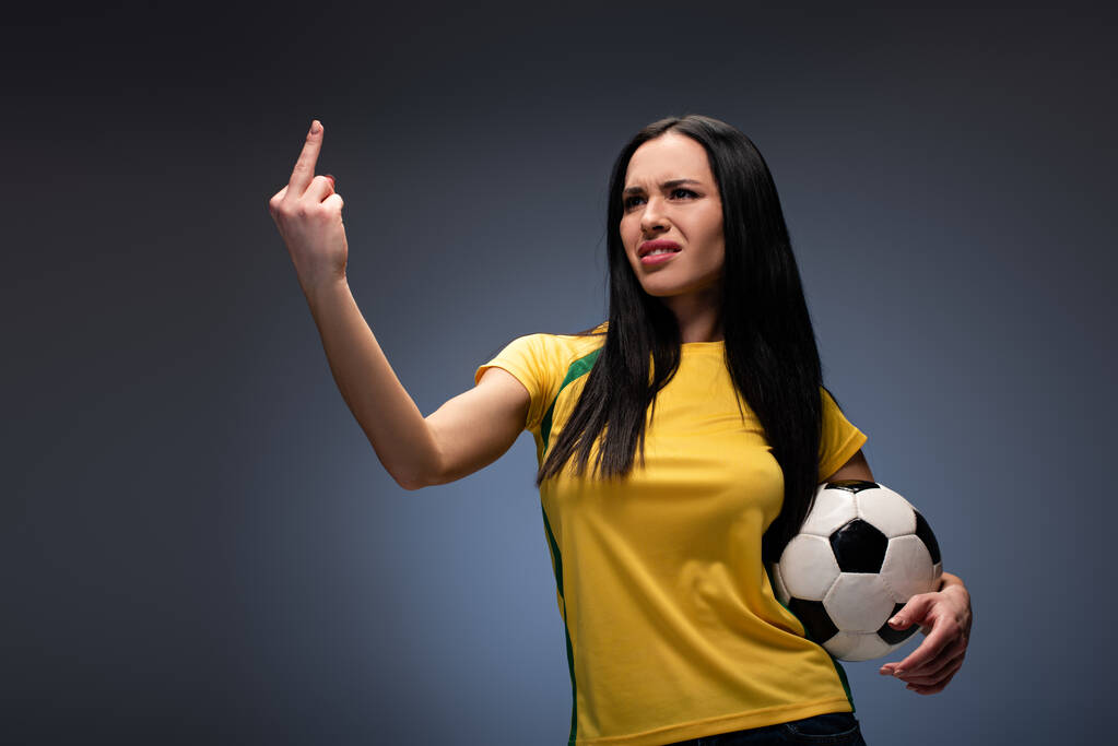 красивый агрессивный футбольный фанат держит мяч, показывая средний палец на сером
 - Фото, изображение