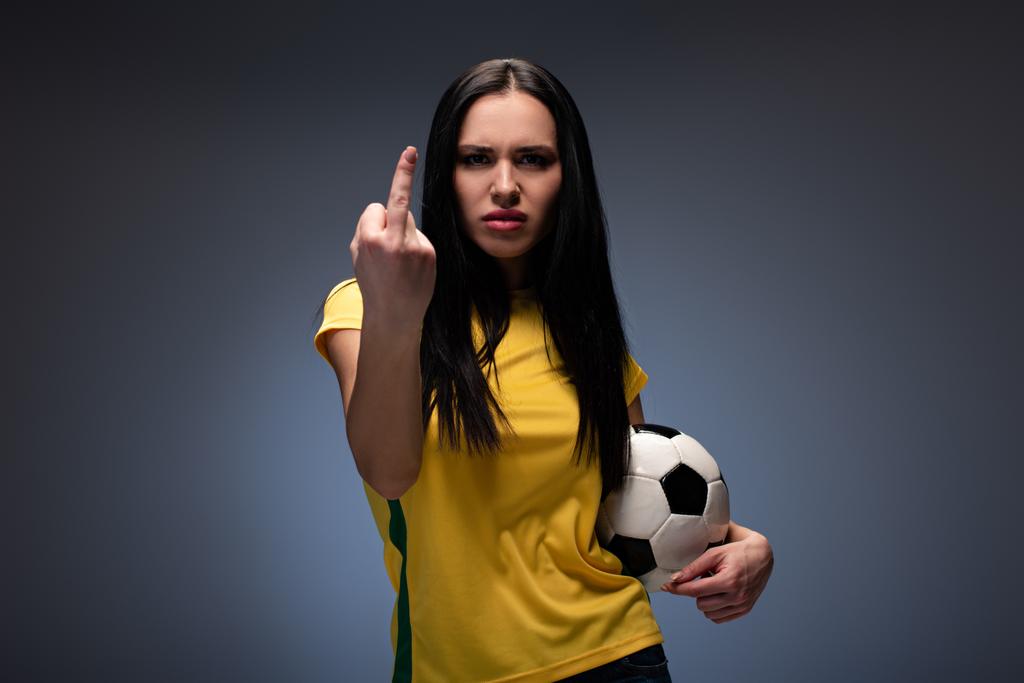 θυμωμένος θηλυκό οπαδός του ποδοσφαίρου κρατώντας μπάλα, ενώ δείχνει μεσαίο δάχτυλο στο γκρι - Φωτογραφία, εικόνα