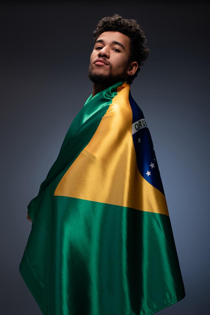 trotse Afrikaanse amerikaanse voetbal fan verpakt in Braziliaanse vlag op grijs - Foto, afbeelding