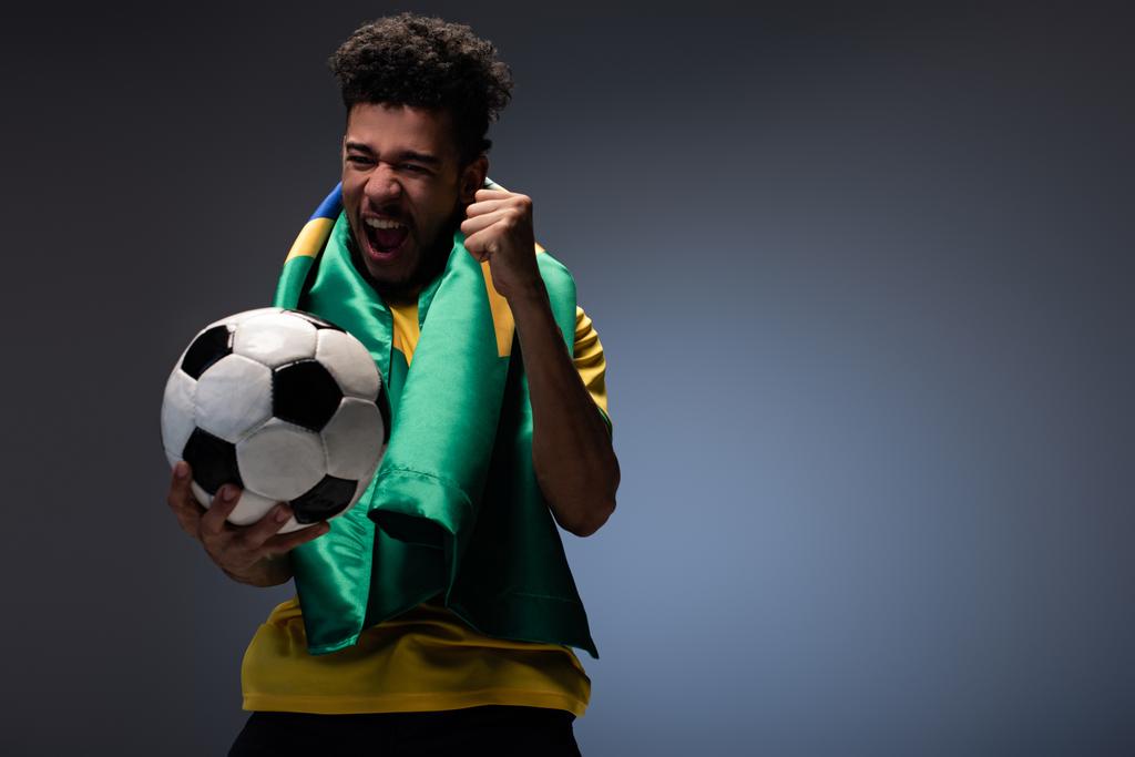 Веселый африканский американец с бразильским флагом, кричащий и держащий футбольный мяч на сером
 - Фото, изображение