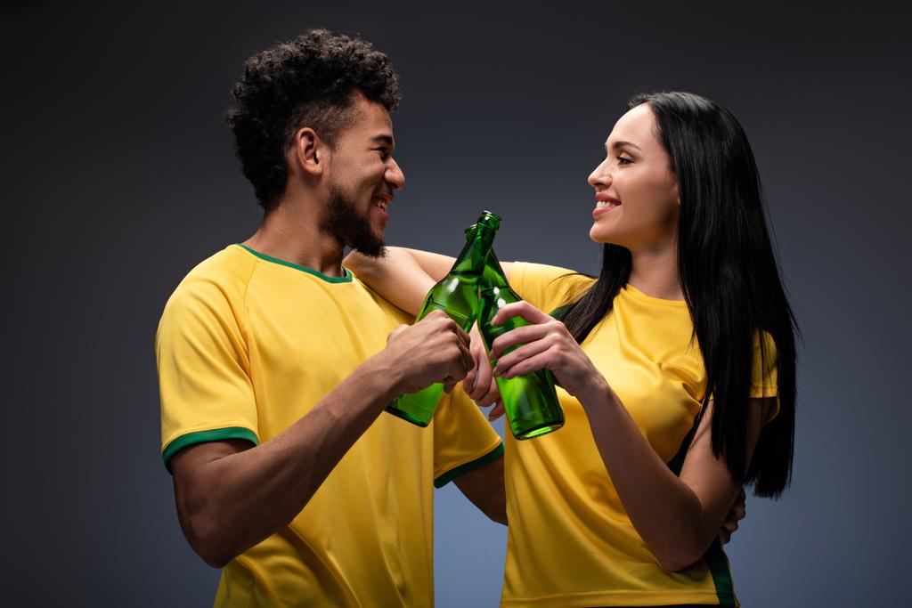 couple multiethnique souriant de fans de football en t-shirts jaunes cliquetis avec des bouteilles de bière sur gris
 - Photo, image