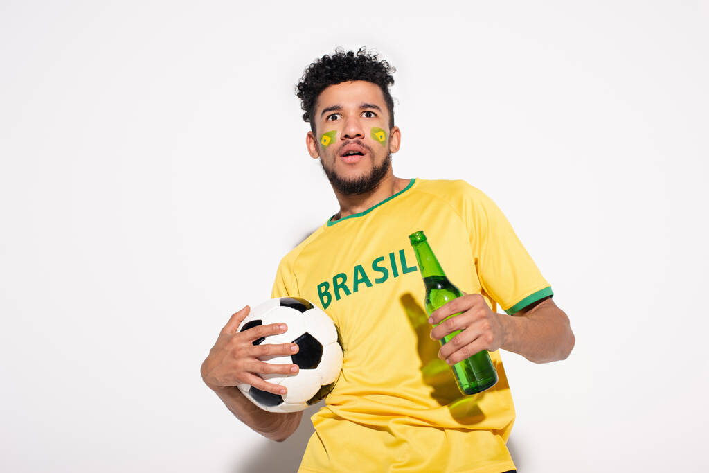 šokovaný africký americký fotbalový fanoušek drží míč a láhev piva na šedé - Fotografie, Obrázek