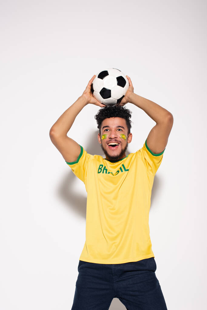 vzrušený africký americký fotbalový fanoušek ve žlutém tričku drží míč na šedé - Fotografie, Obrázek