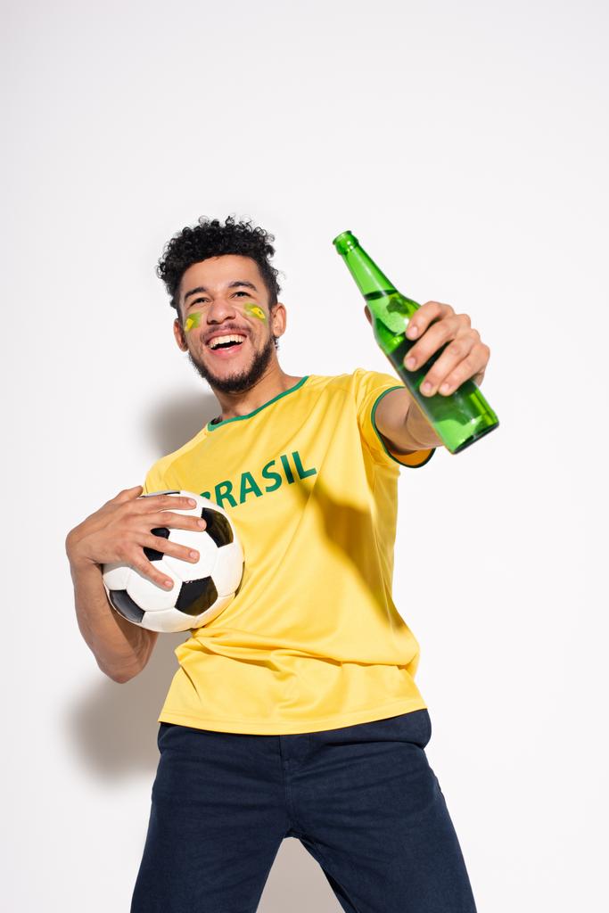 χαμογελαστός Αφροαμερικάνος κρατώντας μπάλα ποδοσφαίρου και ένα μπουκάλι μπύρα στο γκρι - Φωτογραφία, εικόνα