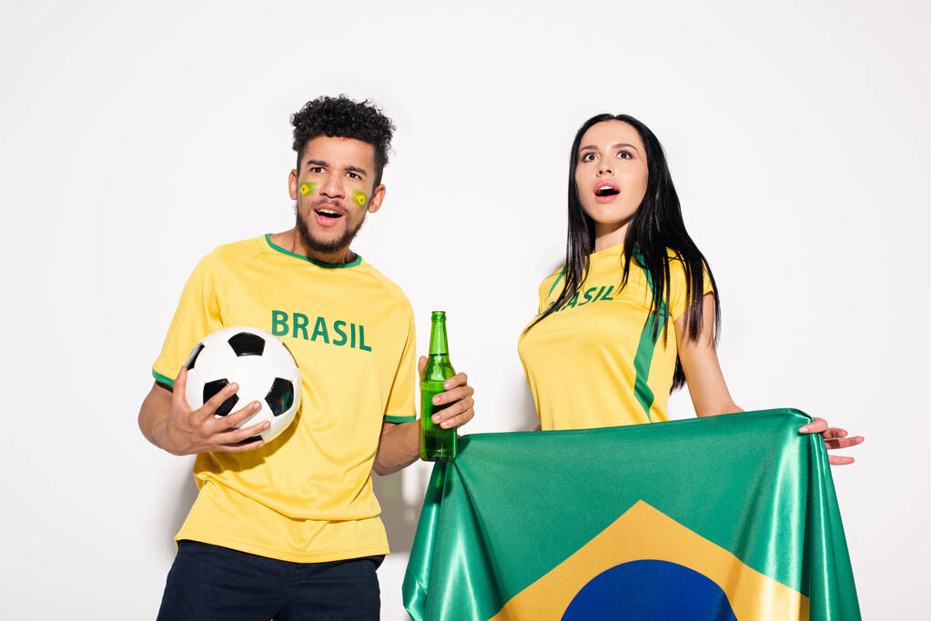 multikulturális pár meglepett labdarúgó rajongók kezében brazil zászló, labda és üveg sör szürke - Fotó, kép