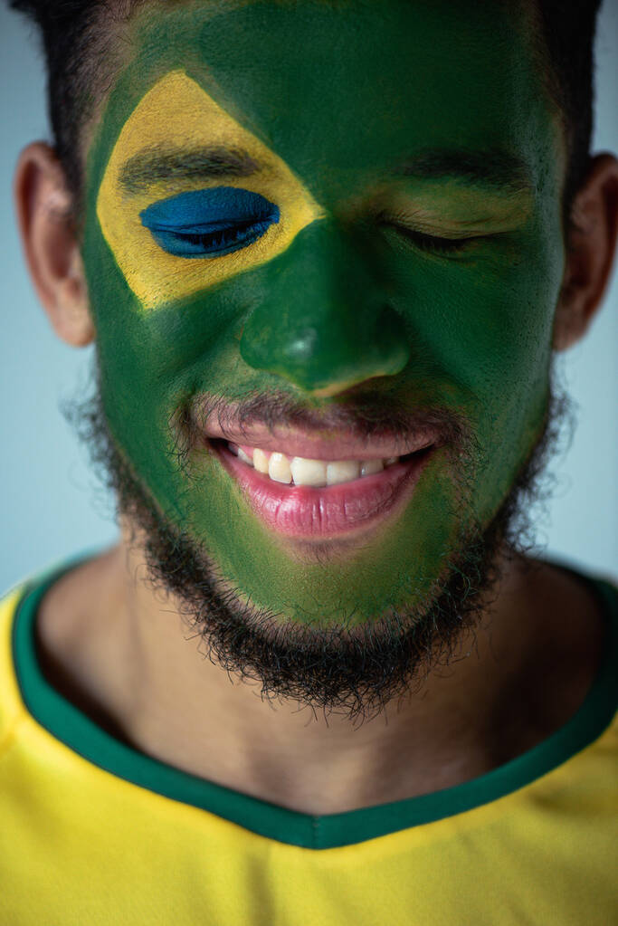sorridente fã de futebol americano africano com olhos fechados e rosto pintado como bandeira brasileira isolada em cinza
 - Foto, Imagem