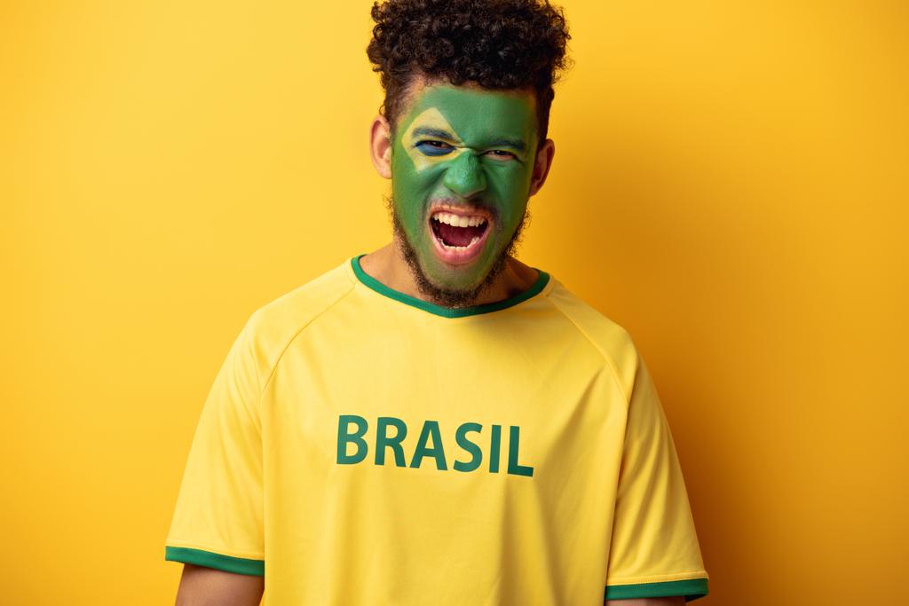 african american football fan with painted face in t-shirt with brazil sign yelling on yellow - Photo, Image