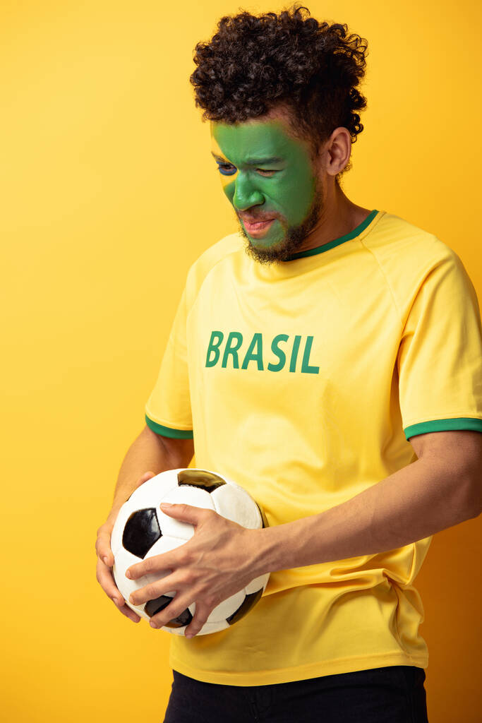 zestresowany amerykański fan piłki nożnej z twarzą namalowaną jako brazylijska flaga trzymająca piłkę na żółto - Zdjęcie, obraz