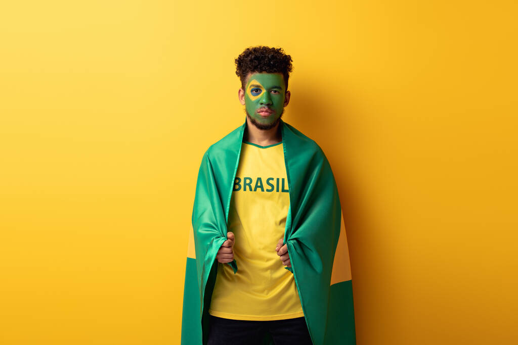 Серйозний афро-американський футбольний фанат з намальованим обличчям, загорнутим у бразільський прапор. - Фото, зображення