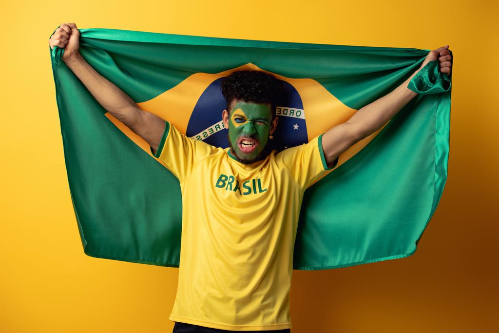 Emotionaler afrikanisch-amerikanischer Fußballfan mit gemaltem Gesicht und brasilianischer Flagge auf gelb - Foto, Bild