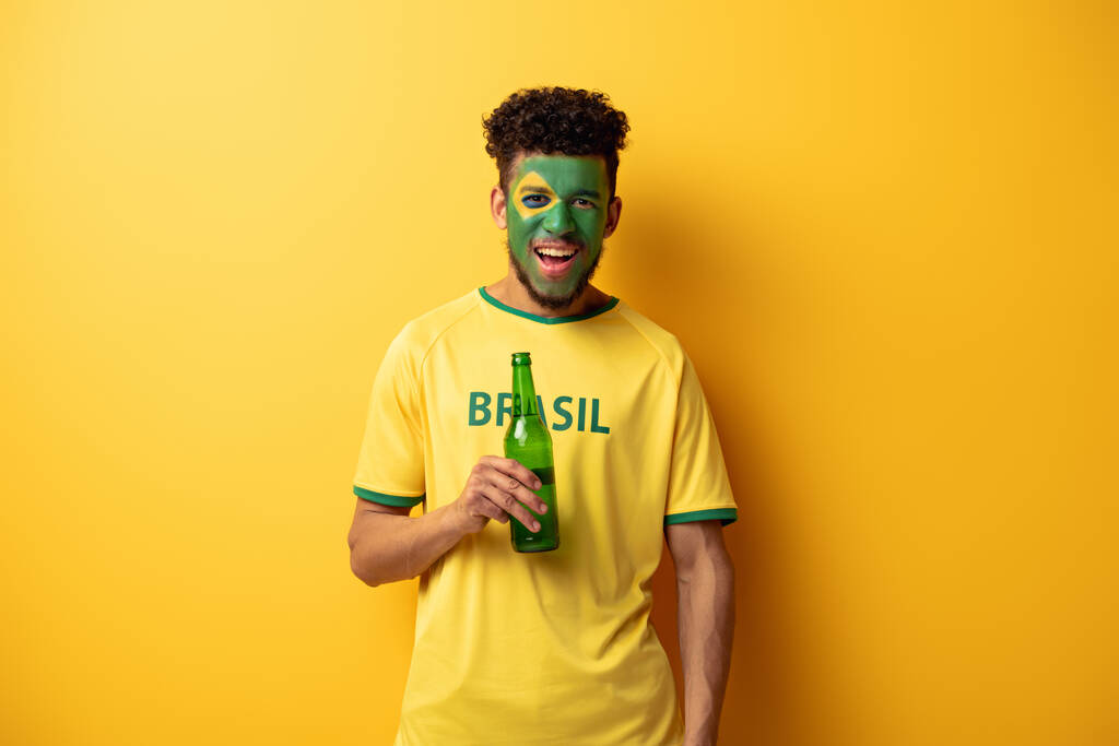 vrolijk afrikaanse amerikaanse voetbal fan met gezicht geschilderd als Braziliaanse vlag met fles bier op geel - Foto, afbeelding