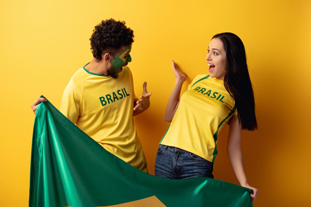 man afrikaanse amerikaanse voetbal fan met geschilderd gezicht en gelukkig meisje gebaren en het houden van Braziliaanse vlag op geel  - Foto, afbeelding