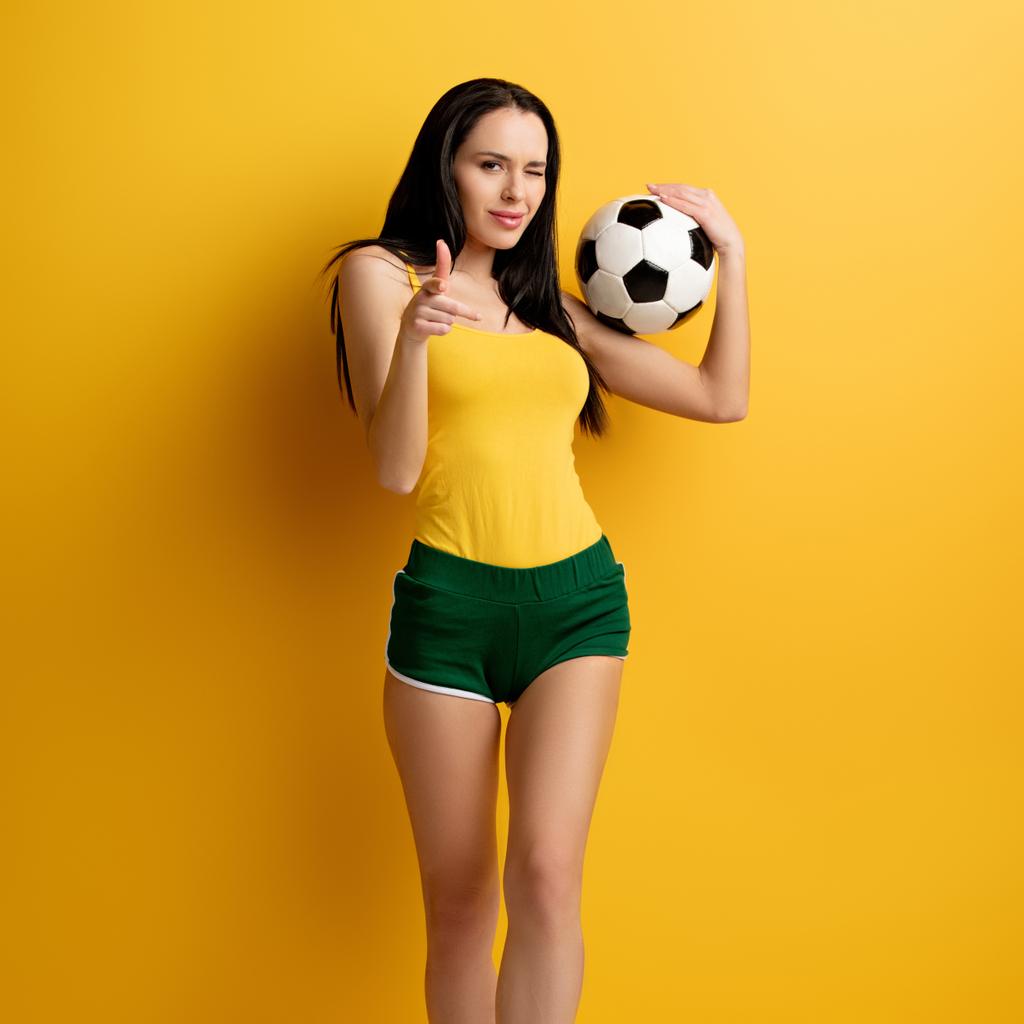 ευτυχισμένη γυναίκα οπαδός του ποδοσφαίρου με μπάλα κλείσιμο του ματιού και δείχνοντας σας στο κίτρινο - Φωτογραφία, εικόνα