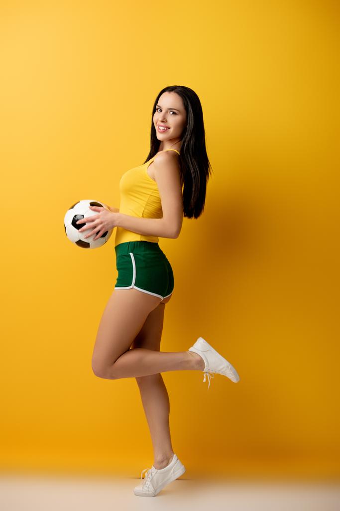 cheerful female football fan in shorts holding ball on yellow - Photo, Image