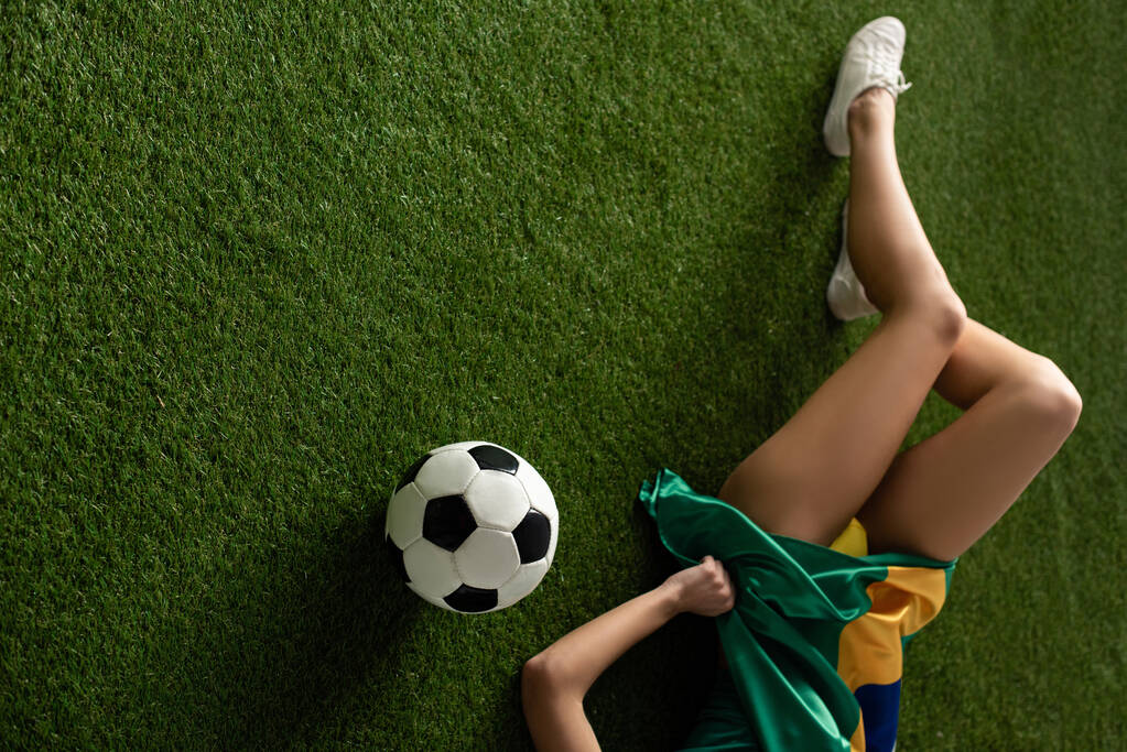 vista cortada de menina sexy deitado com bandeira brasileira e bola de futebol na grama verde
 - Foto, Imagem