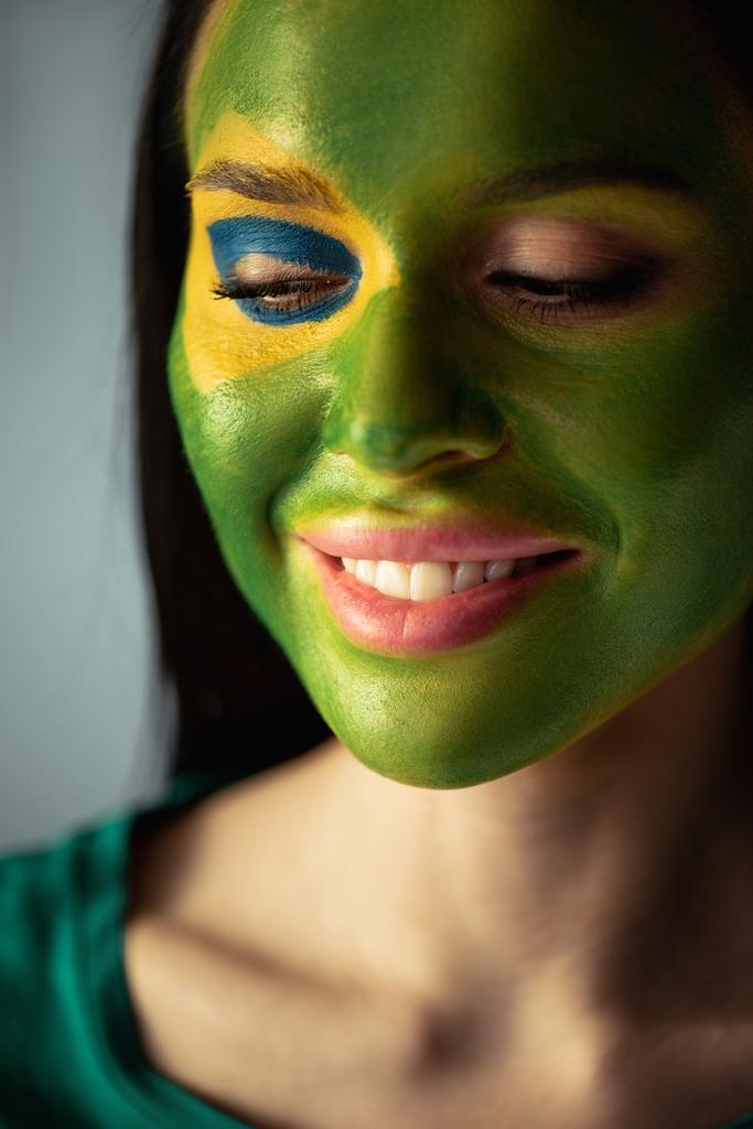 attractive cheerful woman with painted brazilian flag on face  isolated on grey - Photo, Image