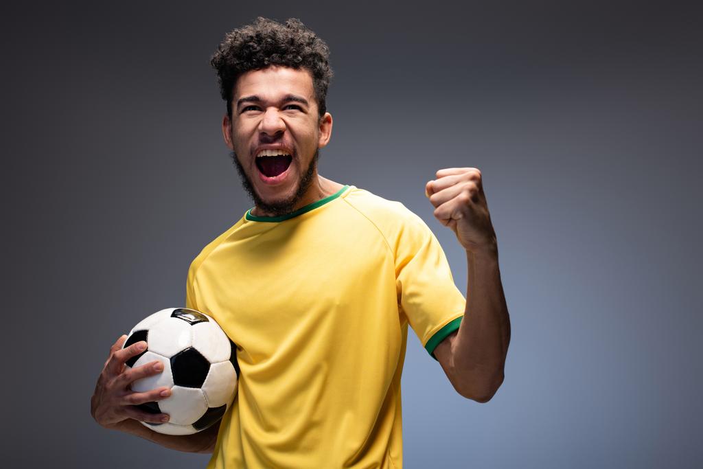 vzrušený muž africký americký fotbalový fanoušek ve žlutém tričku drží míč na šedé - Fotografie, Obrázek