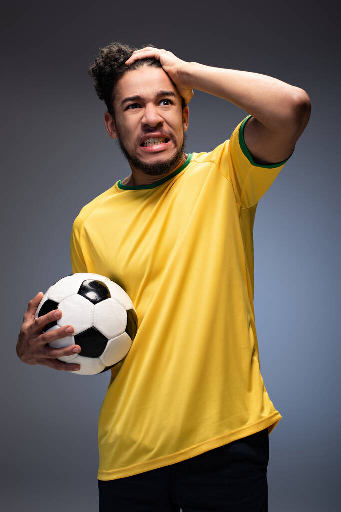 τόνισε αφροαμερικανός ποδοσφαιρόφιλος σε κίτρινο t-shirt κρατώντας μπάλα σε γκρι - Φωτογραφία, εικόνα