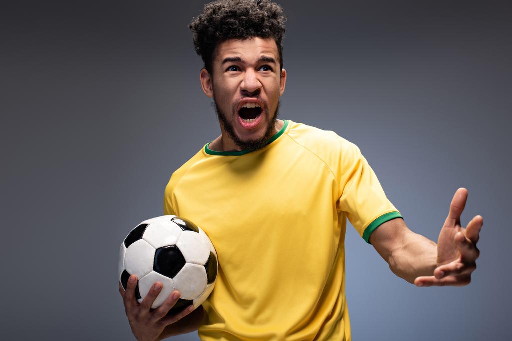 Wütender afrikanisch-amerikanischer Fußballfan in gelbem T-Shirt schreit und hält Ball auf grau - Foto, Bild