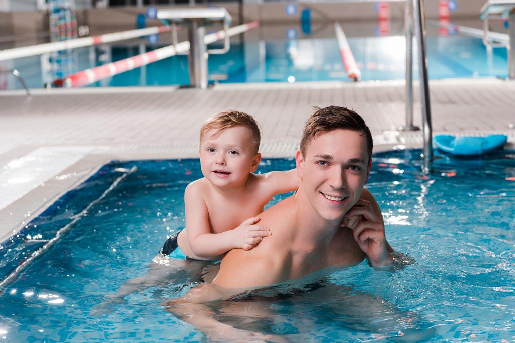 χαρούμενο μικρό παιδί και προπονητής κολύμβησης κοιτάζοντας κάμερα στην πισίνα  - Φωτογραφία, εικόνα