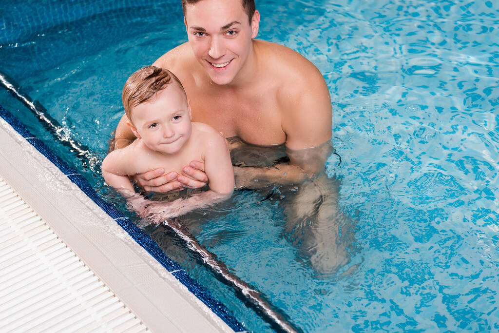 水泳プールで泳いでいる間に腕の中で幼児の男の子を保持する幸せな水泳トレーナー  - 写真・画像