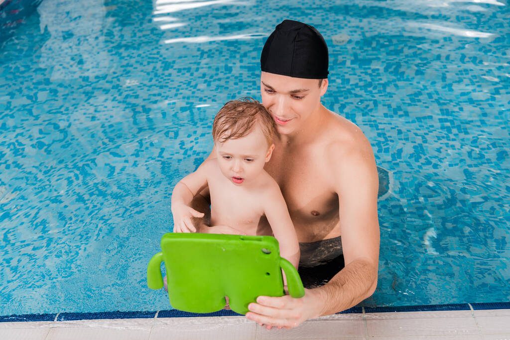 όμορφος προπονητής κολύμβησης και το παιδί αγόρι κολύμπι και κοιτάζοντας ψηφιακή ταμπλέτα  - Φωτογραφία, εικόνα