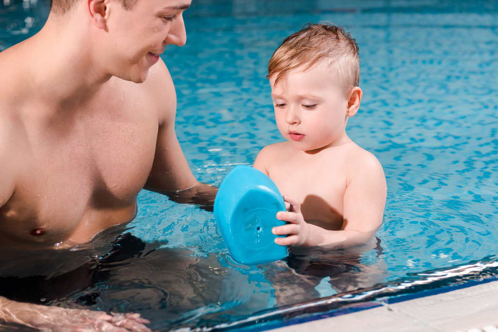 happy swim coach looking at toddler boy playing with toy ship in swimming pool  - Photo, Image