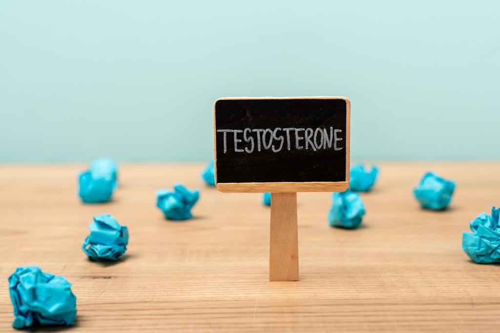Namensschild mit Testosteron-Schriftzug in der Nähe von zerknülltem Papier auf Holzoberfläche isoliert auf blau - Foto, Bild