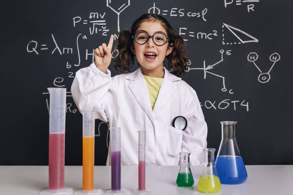 μικρό κορίτσι επιστήμονας με γυαλιά σε εργαστηριακή ποδιά με χημικές φιάλες έχοντας μια ιδέα, σχολείο μαυροπίνακα φόντο με ζωγραφισμένα στο χέρι φόρμουλες της επιστήμης, πίσω στο σχολείο και επιτυχημένη γυναικεία έννοια σταδιοδρομίας - Φωτογραφία, εικόνα