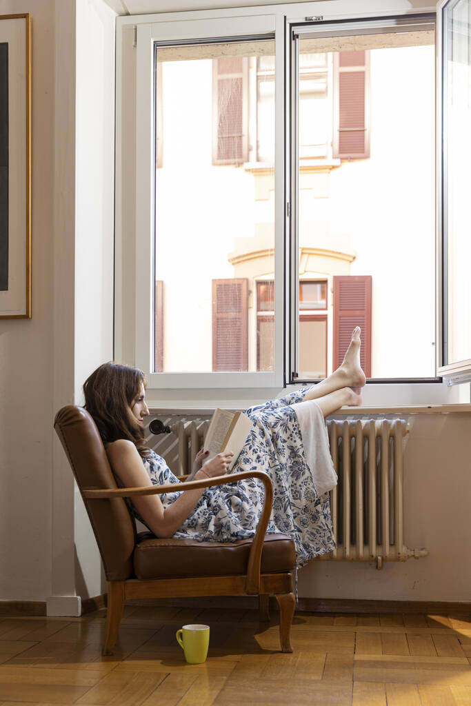 Νεαρό όμορφο κορίτσι με μακρύ φόρεμα κάθεται σε μια παλιά πολυθρόνα και διαβάζοντας ένα βιβλίο - Φωτογραφία, εικόνα