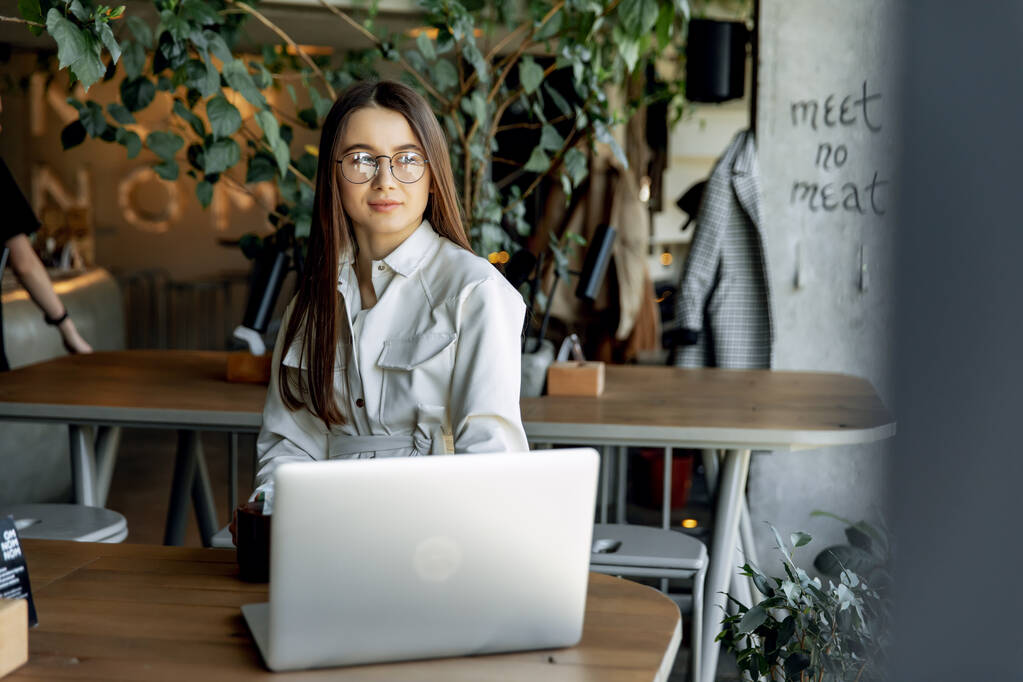 Urocza kobieta freelancer w modnej odzieży pracującej na laptopie podczas porannego śniadania w przytulnej kawiarni. Ładne młode dziewczyny z brązowymi włosami uśmiechnięte i pozujące przed kamerą. - Zdjęcie, obraz