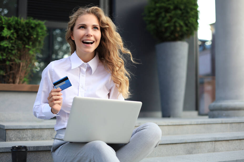 Concepto de compras online. Mujer rubia joven sosteniendo una tarjeta de crédito y haciendo el pago en línea con el ordenador portátil al aire libre
 - Foto, Imagen