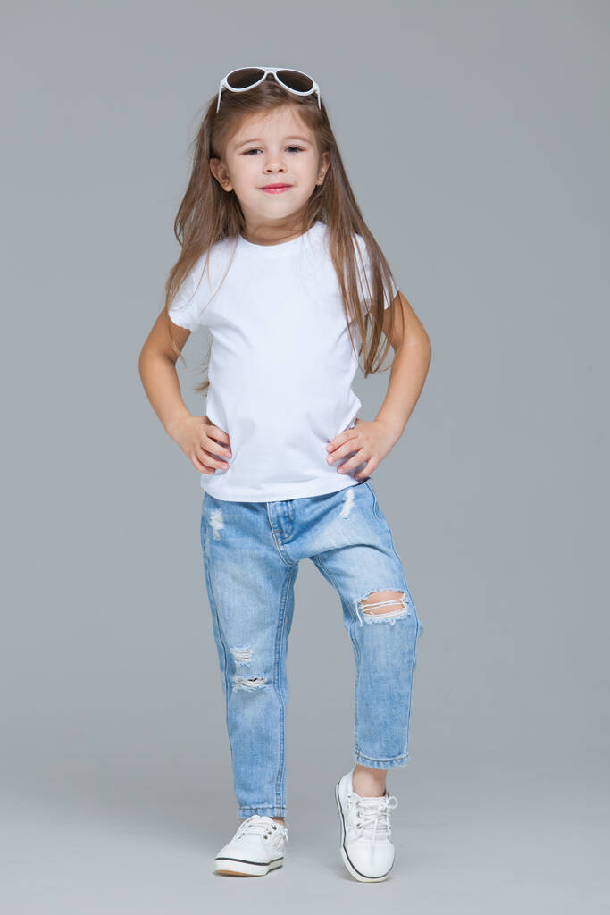 Niña preescolar en jeans azules, camiseta blanca y gafas de sol posando aislada sobre fondo gris
 - Foto, Imagen