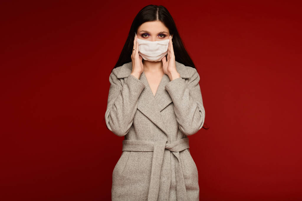Young brunette woman in a protective face mask and coat isolated at the red background. Model girl in medical mask and spring outfit, studio shot with copy space for your text. Anti-flu, anti-epidemic - Zdjęcie, obraz
