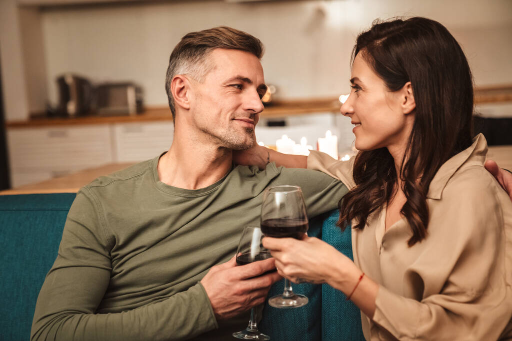 Εικόνα του ευτυχισμένου παθιασμένου ζευγαριού πίνοντας κόκκινο κρασί από ποτήρια ενώ έχοντας ρομαντικό δείπνο υπό το φως των κεριών στο σπίτι - Φωτογραφία, εικόνα