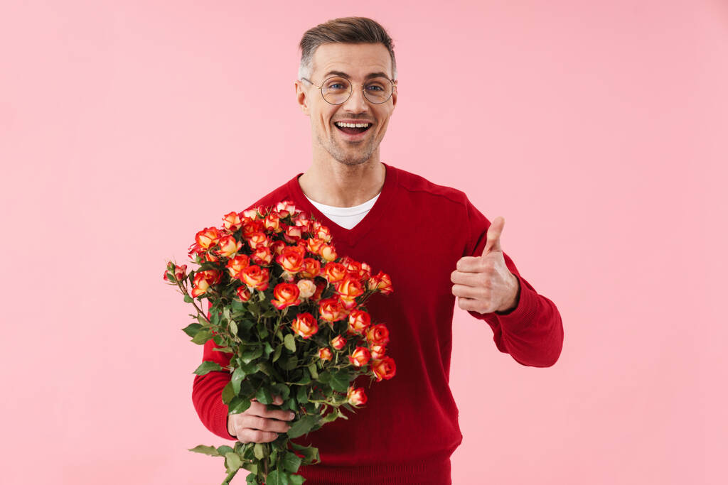 ハンサムな白人男性の肖像画眼鏡をかけて花を保持し、親指を示すピンクの背景に孤立 - 写真・画像